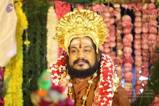 A man seated in a floral setting wears an ornate gold crown with a central deity-like figure, a red-brown saffron-toned shawl, a red and white rose garland, and a gold-toned ceremonial necklace with a rudraksha-style mala. He has a vermilion tilak on his forehead and a full dark beard. Behind him hang strands of pink and white flower garlands and yellow marigold strings, with green foliage on the left. Appearance-based label: a person, seated in person at a celebration, with the page noting Avatar Day festivities.