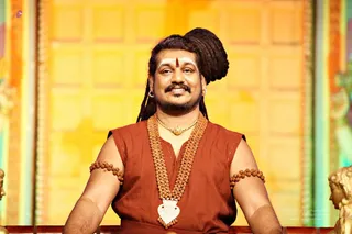 A smiling man with long dreadlocked hair swept up into a top knot sits centred against a warm yellow panelled background. He wears a sleeveless rust-orange garment, multiple rudraksha malas across the chest, rudraksha armbands, a gold necklace with a white heart-shaped pendant, and drop earrings. A red bindi and white vertical tilak mark his forehead. Golden figure statues appear at the frame edges. a person: the long swept-back hair, saffron-orange clothing, rudraksha malas and ceremonial gold necklace are consistent with the page's subject, shown in person on a stage setting.