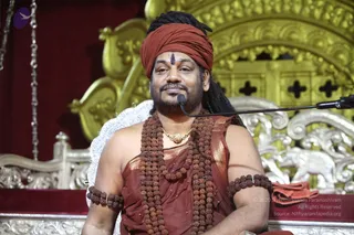 A seated man in a rust-red turban and rust-red draped garment sits before an ornate gold and silver carved throne with a microphone stand in front of him. He wears multiple strands of rudraksha beads, rudraksha armbands, a gold pendant necklace, and a vertical tilak on his forehead. Behind him stands a gold-carved throne backdrop with a red velvet cloth. He is seated and speaking into a microphone.