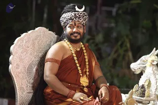 A seated man in an ornate silver-backed throne chair wears a leopard-print turban with a white crescent ornament, a rust-orange draped garment, and a heavy gold bead necklace with a large gold pendant. Red rudraksha-style beaded armlets are on his upper arm and wrist, and he has a vermilion forehead mark and a gold ring. He smiles toward the camera. Beside him is a silver deity-shaped vehicle with a bull-like head and flowers. Dark foliage forms the background.