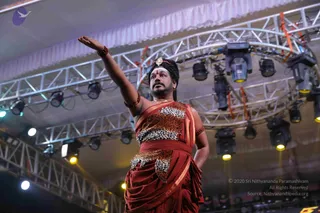 A man stands on a stage under a metal truss rig fitted with stage lights, raising his right arm upward with an open palm. He wears a rust-red dhoti and a red wrap, with leopard-print panels across his chest, waist and draped cloth, and gold-edged borders. He has a red-orange tilak on his forehead, a jewelled ornament at his crown, rudraksha bracelets on his arms and a gold chain at his neck. His hair is long and swept back, and his face is bearded. The setting is a large indoor event hall with white draped ceiling fabric and overhead spotlights.