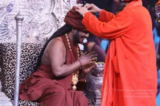 A seated man in a maroon-red robe and dark red turban sits on an ornate silver throne draped with leopard-print fabric, smiling with hands pressed together in a namaste-like gesture. He wears a heavy gold ceremonial necklace and a red wristband. A standing man in an orange-saffron robe and beard reaches forward to place his hands on the turban, appearing to give a blessing or crown-like honour. A silver ornamental pedestal and a silver lion-head sculpture are visible nearby.