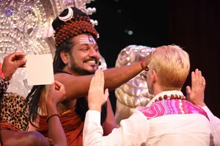 Photograph of a stage event. A long-haired man with a crescent-moon ornament and a tall crown of rudraksha beads, wearing orange-brown clothing and white-and-red forehead markings, smiles while reaching forward and touching the forehead of a blond man in a white shirt with a pink-and-white patterned shawl and a rudraksha mala. The blond man has palms raised in a gesture of reverence. A hand at left holds up a blank white card. Behind them is a silver, sparkling backdrop with a gold lion-like sculpture. Probable Swamiji is the crowned man with long hair, rudraksha crown and orange-brown robes, performing a blessing touch on a devotee.