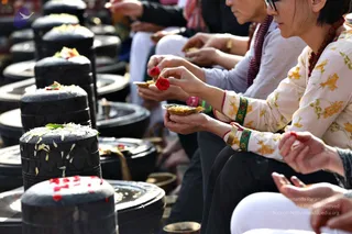 Close photo of seated devotees performing Rudra Abhishekam at rows of black stone lingams on metal bases. In the foreground a woman in a cream floral top with a red-beaded mala and green-and-red bangles holds a small brass dish and a red flower over a lingam. Beside her, another woman in a grey top and red beaded necklace holds a brass dish, and more hands hold offerings, some with red petals and white flowers scattered on the lingams.