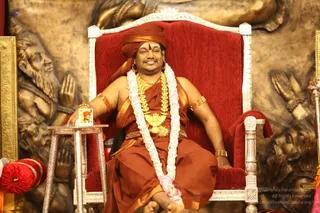 A man sits on an ornate red-upholstered throne with silver-trimmed arms and back, in front of a gold-toned carved relief panel of figures with raised hands. He wears a rust-orange turban, a rust-orange dhoti-style wrap, a heavy gold necklace and a white-and-pink flower garland, with gold armlets and bangles and a red-orange sash across the chest. A small framed photograph stands on a silver side table beside him, and his bare feet rest on the floor. a person: the orange turban, ornate gold ceremonial necklace and garland match the page's a person context, but appearance alone is not conclusive.