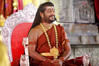 A man with long dreadlocks and a short beard sits on an ornate silver-and-red throne, speaking into a microphone on a black stand. He wears a rust-red wrap over bare shoulders, a heavy gold necklace of round beads with an ornate gold pendant, red beaded armbands, a gold waist belt, and a white and red tilak on his forehead. Behind him stands a carved stone temple wall with red drapery. a person: the long dreadlocks, saffron-red garb, ceremonial gold necklaces and ornate throne match the cues for a person, seated as the teacher giving a discourse.