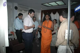Indoor office scene with a conference room sign on the glass door at right. A central figure in an orange robe with a long orange shawl and a long wooden bead mala necklace, with long swept-back dark hair and a red bindi, stands smiling with open hands while speaking to a man in a white shirt and dark trousers. Other people stand nearby, including a man in a light blue shirt, a man in a striped shirt, a woman in a grey patterned sari and a man in a checked shirt at right. Curtains, a wall clock, an air conditioner and a small armchair are in the room. Swamiji (probable): the central orange-robed figure has long swept-back hair, saffron clothing and a rudraksha mala, matching the page's claim.