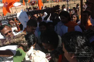 Outdoor festival crowd on dry ground beneath orange saffron flags on poles. Many people press around a central figure with a white flower garland and a pale forehead mark, who holds or touches long wooden poles or carrying staffs. Nearby, a man in white with sunglasses and tilak marks, a young man in an orange saffron shawl with a white forehead mark, and women in saffron and green, plus others in white, crowd closely. A white cloth bundle is held at the lower left. Tent structures and colourful banners appear in the background.