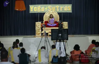 A seated figure in a lavender-purple garment sits cross-legged on an ornate gold throne with a red velvet seat and arched back, centered on a stage beneath a banner reading Yoga Sutras. The seated figure wears a long rudraksha mala and a gold necklace. In the foreground, a row of seated audience members wear white garments with yellow shawls and headphones; one wears an orange robe. Video cameras on tripods and a monitor face the stage, and a dark blue curtain forms the backdrop.