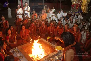 Night photograph of a homa fire ceremony in an open courtyard. A large square brick fire pit blazes at centre, with an officiating priest bare-chested in a dark wrap and long dark hair facing it from the right, pouring from a golden ladle. Around the pit, a large seated circle of devotees in orange and saffron robes, many with white tilak marks and rudraksha malas, sit cross-legged. Several men in orange and saffron stand at the back, and white-clad devotees, some in white saris, line the background. Brass bowls with offerings sit on the low altar in front. A golden deity murti with red and gold decoration stands at upper right.