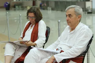 Two seated devotees in white ritual clothing sit side by side in a bright, empty hall with metal stanchions and chain barriers behind them. The woman on the left wears white embroidered kurta and a red-orange shawl with dark patterns, reading a booklet with reading glasses and holding a pen. The man on the right, with short grey hair, wears a white kurta and white trousers, a red-orange shawl draped over his lap, and rests an open notebook on his knee while looking forward. A faint background figure and a dark bin appear behind the barrier. The lower-right corner carries a copyright notice.