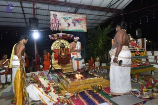 Night photograph of a Shiva Deeksha ritual under a open-sided pavilion with a corrugated roof and string lights. A seated figure in a red-brown robe and rudraksha mala sits on an ornate gold throne with a multicolored canopy, appearing to be the a person, though the appearance is uncertain. Beside the throne, a shaved-headed priest in white dhoti with orange-and-white cloth and a rudraksha mala performs the fire ritual over a raised homa pit with a visible flame. Another bare-chested man in a white dhoti and gold-bordered cloth stands at left holding a garland, and a bare-chested woman at right holds a bell beside a decorated offering table with fruit, brass vessels and marigold garlands. Orange-robed attendants stand in the background. A large poster on the wall shows a robed figure with a map of the world and text in Kannada and English.