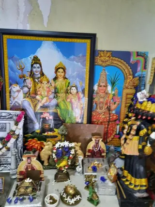 A home shrine photographed head-on. A large framed print on the left shows Shiva seated with Parvati, Ganesha on Shiva's lap and a young Murugan beside them, with a white bull, snowy peaks behind, and a lingam with a lotus at the base. To its right a framed image of a deity holding a sugarcane stalk and a trident-like staff, wearing a tall crown and red garments, stands in a golden arch. In front are small carved figures of seated figures in red robes and a brass lamp, a bowl of white jasmine flowers, a brass kalasha-style vessel, and a garland of pink and white flowers. A dark wooden murti in yellow-trimmed cloth stands at the right edge. No people are present.