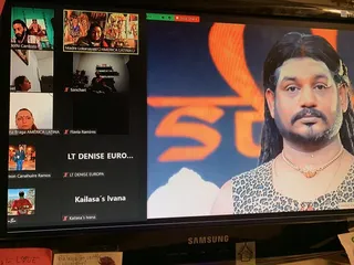 Photograph of a computer monitor showing a video conference grid. On the large right-hand feed, a man with long dark hair, a goatee and a leopard-print sleeveless top with a gold pendant necklace appears against an orange background with an orange symbol. The left-hand grid shows small video tiles of several participants, some with devotional images behind them, a Samsung monitor below, and handwritten notes at the bottom edge. Screen-displayed people are not counted as physical people.