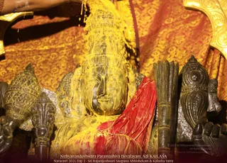 A dark stone deity murti with a tall crown is being bathed with a thick yellow liquid poured from above by a hand entering from the top left, the yellow flowing over the head, face and shoulders. The murti is draped in a red cloth and framed by golden brocade fabric behind it, with dark carved ceremonial objects on either side. The caption along the bottom identifies this as Sri Rajarajeshwari Snapana Abhishekam and Kalasha Yatra on Day 2 of Navaratri 2021 at Nithyanandeshwara Paramashiva Devalayam, Adi KAILASA. No people are fully visible; only a forearm enters the frame.