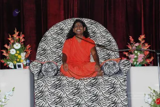 A seated man in long flowing saffron-orange robes sits cross-legged on a large zebra-striped armchair with black-and-white animal print, a red bindi on his forehead, long dark hair swept back, and a beaded chain or mala draped across his lap. Behind him hang deep red velvet curtains flanked by floral arrangements of orange gladioli, white daisies and pink flowers, with a microphone stand to the right. a person is the central figure, seated on a raised stage as a speaker or guru during a satsang-style gathering.