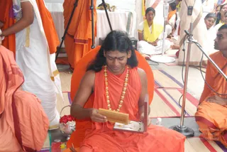 A photo of a man seated cross-legged on an orange cushioned seat, wearing an orange robe, a long gold bead necklace, and a red tilak on his forehead. He has long dark hair and holds an open wooden box with a gold chain inside, looking down at it. Around him are other people in orange and white clothes, a microphone stand, a tripod, and a tray with red flowers. A person in a yellow-and-white garment sits in the background.