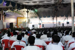A large crowd of seated attendees in white clothing fills a red-chaired hall under a tented canopy with blue-and-white scalloped trim and hanging fans. Their backs face the camera as they watch a stage at the far end. On the stage, a figure in orange robes stands near a small table and a backdrop with a purple banner reading Nithyananda Spurana, with a Pratyaksha Paada Pooja label visible. Speakers, stage lights and a large circular patterned panel frame the scene.
