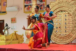 Three young women perform a classical Indian dance on a red stage in a temple-style hall with a golden ornate panel behind them. The dancer in front wears an orange and magenta silk sari with gold border and kneels with one hand raised in a mudra, while the two behind wear blue and pink or orange and blue silk costumes, gold jewellery, garlands and flowers in their hair. A golden palanquin-like chariot and a small deity murti stand at the left, a wall-mounted digital clock shows the time, and framed photographs hang on the walls.