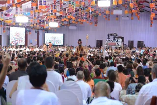 Large indoor satsang hall with a dense crowd of devotees seated on white chairs, many in white and orange clothing, some raising hands. A figure in orange robes with a red and gold turban, wearing a long rudraksha-style mala, sits elevated on a golden seat holding a tall golden staff, with a lit stage. Hanging colourful tasselled lanterns decorate the ceiling, and two large screens show crowds. A camera on a tripod stands at the right side. The room shows devotees gathered around a central guru figure, a person seated on the dais.