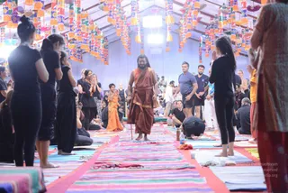 A large tent hall decorated with rows of colourful hanging pennants in red, yellow, orange and blue. A bearded man in an orange-saffron dhoti and shawl, with long dark hair, walks down a central aisle lined with striped rugs and mats. He carries a wooden staff. Around him, participants in black clothing stand with palms pressed together in namaskar, some sit on mats, and a man in a grey t-shirt and dark shorts watches. A woman in orange sari stands with hands folded, and one person bends forward in prostration on the mat.