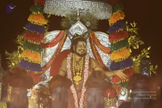A smiling man with long dark hair and a red tilak on his forehead sits on an ornate silver throne under a silver canopy, decorated with multicoloured tassels in red, orange, green, blue and purple, peacock feathers and gold foliage. He wears a pink and white floral garland, layered gold necklaces with a large gold pendant, red and gold drapes, and extends one arm outward. In the foreground, several people in dark clothing and a figure in a head wrap are partly visible. a person is seated on the throne in saffron-red robes with gold jewellery and a garland, and the scene is lit warmly against a dark background.