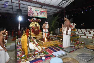 Evening ritual in an open-sided pavilion with a corrugated roof and string lights. A seated man in an ornate golden-cushioned throne wears a red-orange garment and beaded necklaces, flanked by a red canopy. Beside a square gold-toned fire pit with a lit flame, a bare-chested priest in a white dhoti and pink-white head cloth performs the rite. A standing man in white dhoti with a gold necklace holds his hands together near the flame. Brass vessels, flower garlands, banana leaves and fruit surround an altar with a draped deity murti in saffron. Several robed attendants in orange and saffron stand in the background. A wall banner shows a map with a framed portrait labelled text. Homa fire ritual setting.