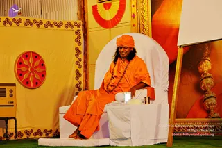 A man in saffron-orange robes and an orange turban sits on a large white throne-style chair on a green carpeted stage. He wears a long rudraksha mala and smiles with eyes lowered, resting his hand on the armrest. On the white table beside him are a small lamp, a white cloth-wrapped item and a metal tumbler. Behind him hang a red-and-yellow fan emblem, a cream backdrop with a red sun-wheel motif and a large framed image of a golden urn-like finial. Wall-mounted air cooler at left. A seated person in saffron on a throne, presented as a discourse or satsang setting.