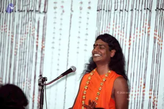 A smiling man with long dark hair and a red bindi stands at a microphone on a stand, speaking or singing. He wears a sleeveless saffron-orange robe and a long ceremonial necklace of gold beads. His hands are raised in front of him. Behind him hangs a pale backdrop covered with vertical strands of beads in red and dark tones. One other person's dark head is partly visible at the lower left edge.
