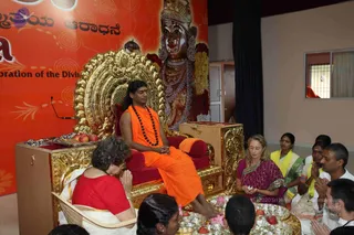 A seated man in an orange saffron robe with a rudraksha mala and a red tilak sits on an ornate gold throne with red cushions, with bare feet. Behind him is a large painted deity murti with a crown and garlands, against an orange banner. A woman in a red blouse and white sari sits in front with hands joined in namaskar, while a woman in a magenta sari holds a small offering. Trays with apples, flowers and small brass lamps are arranged before the throne, and devotees in white and yellow sit to the right. a person appears in person on the throne, identified by orange saffron robes, a rudraksha mala and a red tilak.