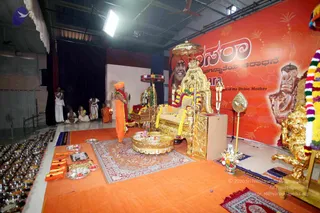 A ceremonial Dasara celebration on an orange-carpeted stage with a large red banner reading Dasara in English and Kannada script. A man in saffron robes with a saffron turban stands on the orange mat before an ornate gold throne with garlands. On the banner, a portrait labelled with a name shows a smiling man in an orange turban beside a golden throne. Rows of small silver vessels with offerings line the left foreground, and a gold umbrella and ceremonial fan sit by the throne. Behind, several people in white dhotis sit and stand near a black curtain, one in orange. A gold peetha-style chair and floral garlands are arranged around the stage.