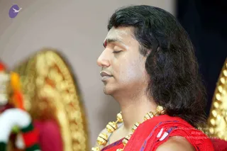 Close profile photograph of a man with long, swept-back dark hair, eyes closed in meditation, wearing a red shawl with a blue-and-white edge over a ceremonial gold bead necklace. A red mark and white paste mark the forehead. Blurred gold ornate ceremonial structure and a red-and-white garland appear in the background. Shawl and gold necklace are worn by the man in the foreground; the gold arch is a background object.
