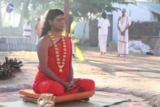 A man with long dark hair sits cross-legged in meditation on a low orange cushion placed on a woven mat outdoors. He wears a red saffron-coloured wrap and a heavy gold necklace of large beads, with a red tilak mark on his forehead, and his eyes are closed with hands resting in his lap. In front of him sits a small brass vessel. Behind him, several people in white dhotis and white shirts stand on a sandy ground near a low block wall, a yellow cloth hangs on a fence, and a green tree and palm fronds frame the morning sunlight. The appearance of the seated man is a probable match for Swamiji (probable), seated in meditation, with the long hair, gold beads and saffron wrap as the qualifying cues.