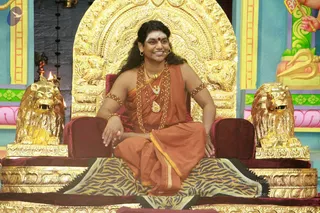 A photograph of a seated man on an ornate gold throne with carved lion-head armrests, flanked by two golden lion statues and a tiger-striped throne cloth. He wears a saffron-orange garment draped over one shoulder, multiple rudraksha malas, a gold necklace with pendant, gold hoop earrings, a red bindi and white forehead markings, and smiles while seated with his hands resting on his knees. Behind him are painted pink, green and yellow panels, and a small oil lamp burns at the left edge. The figure matches the Swamiji (probable) appearance cues of long swept-back hair, saffron clothing, rudraksha malas and ceremonial gold necklace, seated on a throne during a satsang setting.