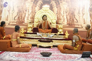 A seated man in saffron-orange robes with a rudraksha mala and ornate gold necklace sits on a large gilded throne with lion armrests and a tiger-skin cover, gesturing with one raised hand during a satsang. Four devotees in orange robes with yellow borders sit on cushions around him: two women at left, and two men at right, one bald with white tripundra markings on his arms and forehead. A red patterned carpet covers the floor, with a brass bowl in front. The backdrop is a carved temple-pillar wall relief. Swamiji (probable): the seated central figure has long swept-back hair, saffron robes, a rudraksha mala and gold necklace, consistent with the page's description of a satsang discourse.
