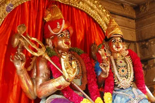Golden metal murti of Shiva and Parvati seated side by side before a red satin backdrop, decorated with pink and red flower garlands and green leaf garlands. The left figure holds a trident and a damaru-like drum with a snake, wears a red turban with a pearl band and rudraksha beads, and has a gold-framed ornate arch behind. The right figure wears a jewelled golden crown, heavy gem-studded necklaces, blue and gold garments and a lotus-like sceptre. Stone carved walls frame the scene. No physical people are visible; the figures are statues.