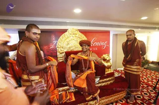 Photo at an indoor event with a red backdrop reading "...kening ...WAR" (partially visible. A central figure seated on an ornate gold-and-red throne with gold lion armrests wears a maroon-red turban, a long rudraksha mala and a ceremonial gold necklace, smiling and holding an orange shawl. Two attendants stand beside him: one on the left in maroon-and-saffron robes with a white tilak and an orange cloth draped over his arm, and one on the right in maroon robes with a rudraksha mala, hands clasped behind. A partially visible bald figure in orange appears at the far left edge. The red-and-white patterned carpet and a potted plant complete the hall setting. Appearance cues (turban, rudraksha, ornate gold necklace, seated on a throne) are present, so the central figure is a probable candidate.