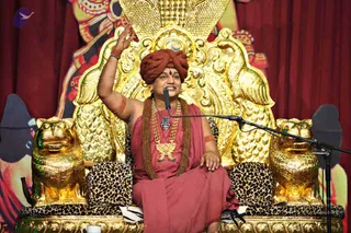 A seated man in a rust-red turban and rust-red robe speaks into a microphone on a stand, raising his right hand. He wears layered gold necklaces, a rudraksha mala, and red-orange bangles. He sits on an ornate gold throne with carved peacock-style backrest and golden lion-head armrests, draped with leopard-print cushions. Behind him hang red curtains and a large painted backdrop. A small white cup sits at the bottom edge. Swamiji (probable): the seated speaker's turban, rudraksha mala, gold necklaces and ornate throne match the page's identification, and the page states he is present.