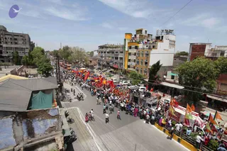 Aerial view of a busy city street during a procession. A large crowd of people, many in white and some in red or orange clothing, walks down the middle of the road. Many red and orange flags and banners are raised above them, and a decorated vehicle with a canopy is visible near the front of the crowd. Multi-storey buildings with balconies line the street, along with trees, parked cars, buses, motorcycles and small roadside shops with awnings.