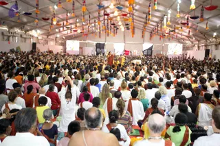 A large photograph of a crowded tented hall filled with hundreds of seated devotees on red mats, many in white and orange-saffron shawls and dresses. Colourful cloth streamers and paper lanterns hang from the tent ceiling. At the front, a person in an orange-brown robe stands on a raised stage with arms outstretched, addressing the assembly, while a golden throne-like seat and dark backdrops are visible behind. Many attendees raise their hands in the air. The scene conveys a large satsang or discourse gathering.