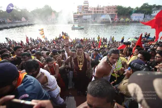 A dense crowd gathers on the steps of a riverside ghat beside a large lake or tank. In the centre, a man with a raised right hand wears a dark maroon-brown robe, a heavy gold necklace and a rudraksha-style mala, walking with a crowd of devotees. Beside him, a bare-chested man wears a white dhoti and a beaded mala, and other devotees wear orange and red garments and white dhotis. Many people hold red and orange flags, and photographers with cameras press in from the right. Behind them are pink temple buildings, a fountain spray over the water and a small boat on the lake.