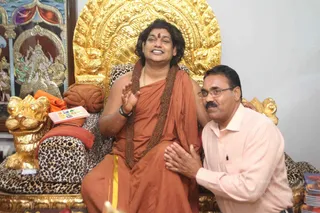 A seated man in saffron-orange robes with a rudraksha mala draped over his shoulders and a red bindi on his forehead sits on a golden, ornate throne with a leopard-print cushion and a gilded lion-head armrest. He gestures with raised right hand, smiling. A man in a light pink shirt and glasses kneels beside him with palms joined in respectful greeting. Behind them is a painted panel of a deity and a framed picture; a book sits on the cushion.