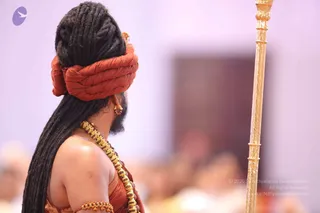 Photograph of a figure seen from the side and slightly behind, with long dark matted locks wrapped up under a rust-orange turban. The figure wears a rust-orange garment, a heavy gold beaded necklace, a gold armlet and a gold ear ornament. A tall gold staff stands on the right edge of the frame. The background is a blurred lavender hall with out-of-focus seated people and a dark doorway.