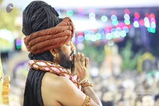 A man in profile with eyes closed and palms pressed together in namaste, wearing a rust-brown wrapped turban with a gold ornament, a long dark braided hair mass, a rudraksha mala with pink flower petals, a gold bangle and gold ring, and bare shoulders. Blurred colourful bokeh lights and a crowd fill the background. Probable Swamiji by appearance: long dark hair, distinctive rudraksha and flower garland, ornate turban; he is physically present, appearing to be in prayer.