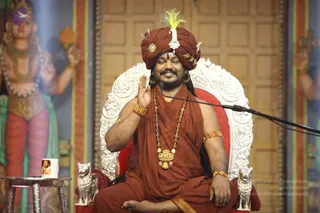 A seated man on an ornate white and red silver-trimmed throne raises one hand in blessing while smiling with eyes closed. He wears a rust-orange turban with a green and white plume and silver ornament, a rust-orange shawl over the shoulders, a long gold necklace with pendant, gold bangles and a red and gold wrist band. A microphone on a stand is at right. Flanking him are silver lion figures, a small side table holds a framed photo mug, and painted deity figures stand on the left and right walls. The setting is a satsang hall. Swamiji (probable): the seated man in saffron-orange turban and ornate gold necklace is presiding as the speaker at this satsang.