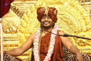 A smiling man seated on an ornate gold throne with carved relief patterns, wearing a rust-orange turban with a gold brooch and feather, a rust-orange wrap over bare shoulders, a gold chain necklace and a long garland of pink flowers. He sits on a leopard-print cushion, arms spread wide, with a microphone on a stand beside him. A red curtain hangs behind.