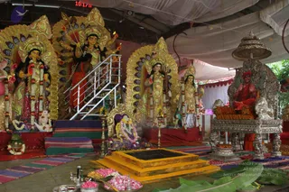 A temple shrine set up under a white tent canopy with several large decorated murtis of goddesses with golden halos and jeweled ornaments, garlanded with marigold and rose strands. The central multi-armed deity murti holds weapons, and a stepped metal ladder leads up toward it. In the foreground stand a brass oil lamp, trays of pink rose petals and offerings on banana leaves, and a yellow stepped platform holding a square bowl. On the right, an ornate silver throne holds a seated figure in red robes and a red turban, while devotees in saffron-orange clothing stand nearby. The scene appears to be a Navaratri darshan at Adhi Kailasa, with no visible fire or abhishekam in progress.