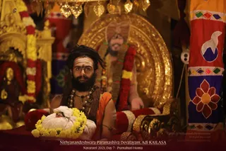 A bearded man with long matted hair and a white tilak on his forehead sits before a golden throne-like backdrop, wearing an orange cloth over his shoulder, rudraksha malas and a red-and-gold garland. He holds a white cloth bundle wrapped with a yellow marigold garland in front of him. Behind him stands a golden murti of a seated figure adorned with a red and yellow flower garland, and a red and blue ceremonial banner with floral motifs hangs on the right, with hanging gold ornaments above. The caption reads Nithyanandeshwara Paramashiva Devalayam, Adi KAILASA, Navarati 2021, Day 7 - Purnahuti Homa. a person is seated in a ritual setting, with ceremonial decor around him.