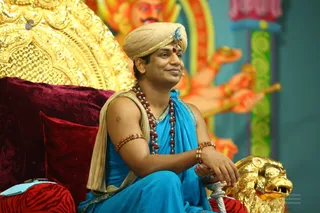 A seated man in a cream turban with a small red bindi and blue accent on the turban sits on a red velvet cushioned throne with a gold-decorated backrest and a golden lion armrest. He wears a bright blue draped robe with gold border, a tan shawl over one shoulder, layered red rudraksha malas and matching rudraksha bracelets, and smiles while resting his hand on the lion. Behind him a large painted deity murti with multiple raised arms holding ritual objects stands against a teal and green backdrop.