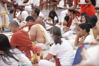 Seated devotees fill a hall during a ritual session. Several men wear white dhotis with gold borders and bare upper bodies, one with a red-orange shawl draped over his shoulders. Women wear white and orange-red saris and rudraksha malas. Some hold small white-and-gold trays or framed items, and a few sit with cupped hands near small gold-decorated objects on the floor. A woman in a white top in the foreground wears a headset. The scene shows a group of about 20 people, with more partially visible at the edges.